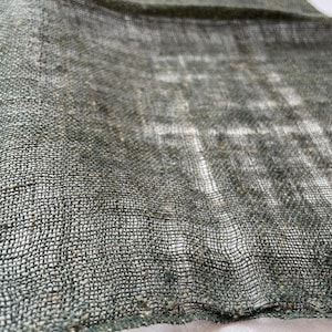 May include: Close-up of a loosely woven, olive green fabric. The texture is visible, with a grid-like pattern created by the interwoven threads. The fabric appears to be lightweight and slightly translucent, with a white background visible through the weave.