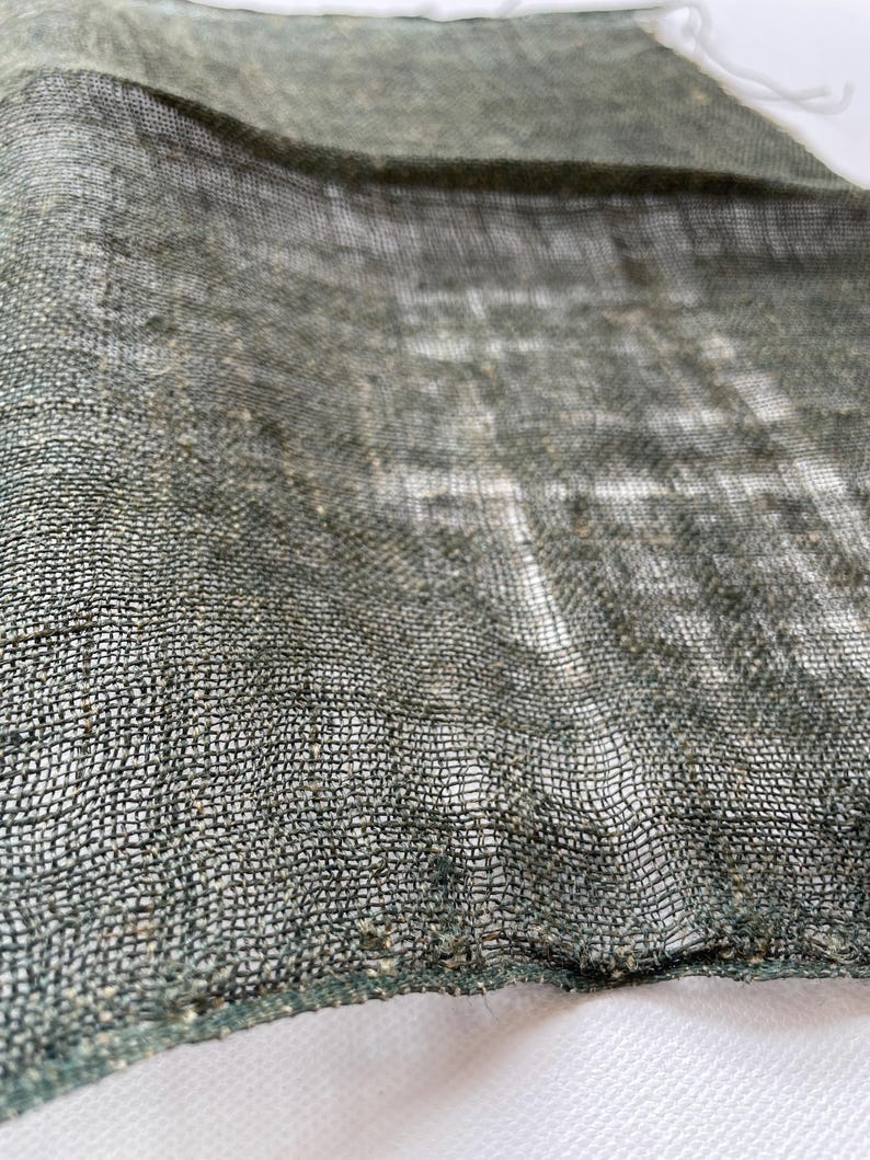 May include: Close-up of a loosely woven, olive green fabric. The texture is visible, with a grid-like pattern created by the interwoven threads. The fabric appears to be lightweight and slightly translucent, with a white background visible through the weave.
