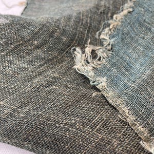May include: Close-up of two pieces of woven fabric with a textured appearance. The fabric is primarily a muted gray-green color, with areas of blue and frayed edges. The weave is visible, creating a grid-like pattern.