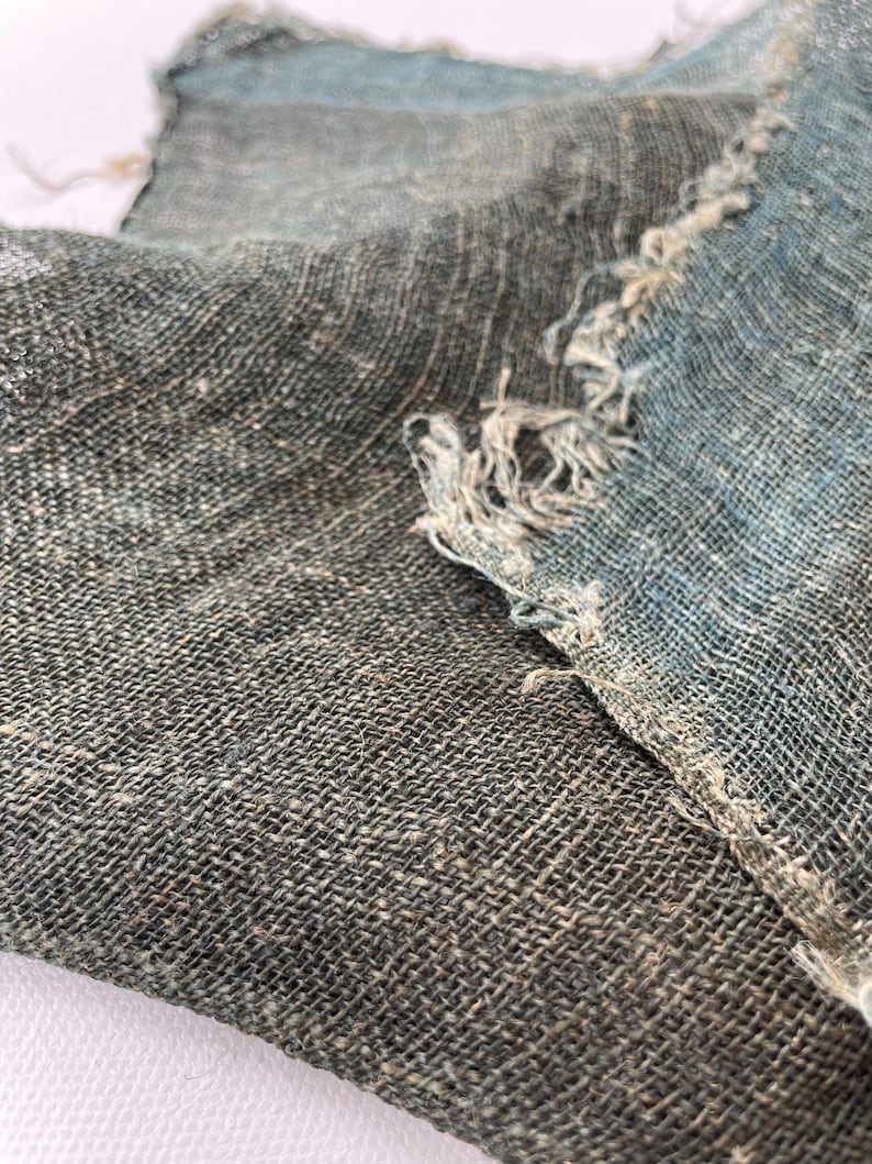 May include: Close-up of two pieces of woven fabric with a textured appearance. The fabric is primarily a muted gray-green color, with areas of blue and frayed edges. The weave is visible, creating a grid-like pattern.