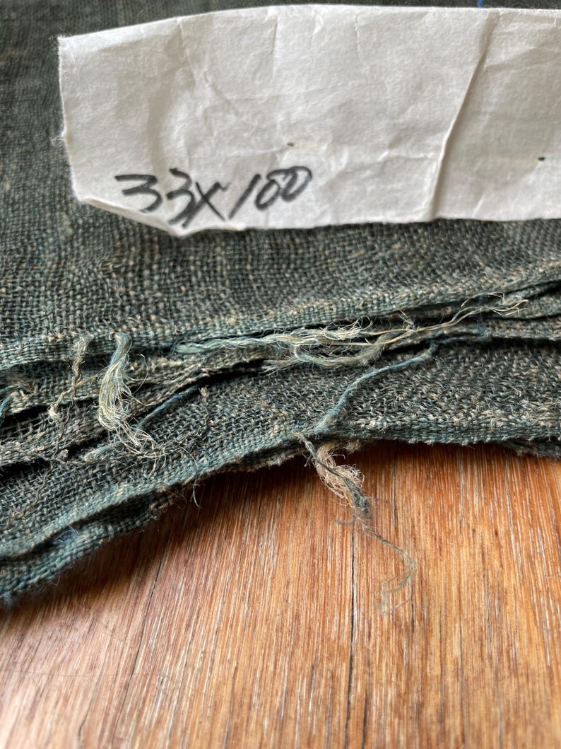 May include: Close-up of a folded, textured fabric in shades of teal and gray. The fabric has frayed edges and a handwritten note that reads "33 x 100". The fabric is resting on a wooden surface.