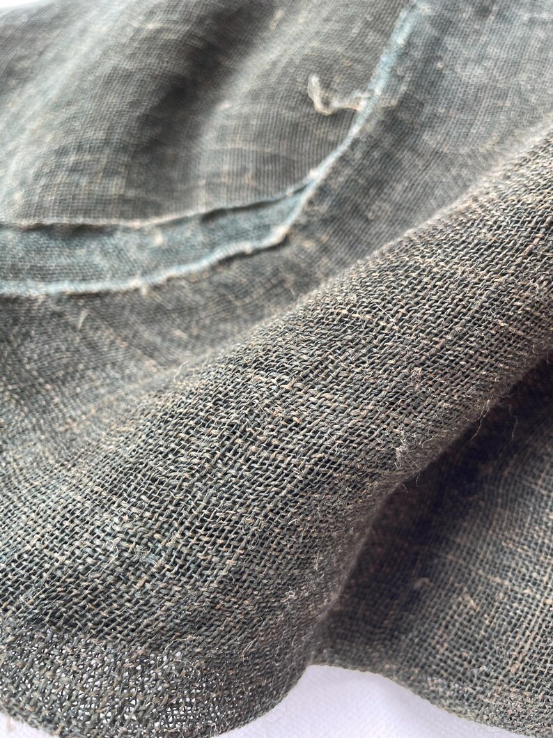 May include: Close-up of a textured, dark green fabric with a loose weave. The fabric appears to be linen or a similar natural fiber, with visible imperfections and a slightly frayed edge. The weave creates a grid-like pattern.