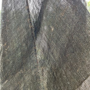 May include: Close-up of a dark gray, loosely woven fabric. The fabric has a textured appearance with visible threads and a slightly frayed edge. The weave creates a grid-like pattern, and the material appears to be lightweight.