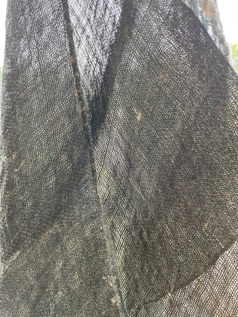 May include: Close-up of a dark gray, loosely woven fabric. The fabric has a textured appearance with visible threads and a slightly frayed edge. The weave creates a grid-like pattern, and the material appears to be lightweight.