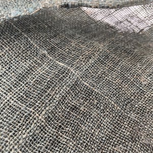 May include: Close-up of a textured, woven fabric in shades of gray and blue. The coarse material has a visible grid pattern, with frayed edges and a slightly open weave. The fabric appears to be natural, possibly linen or burlap.
