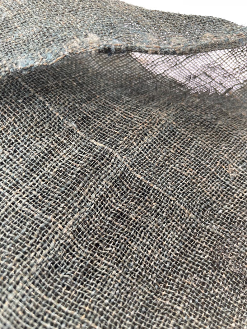 May include: Close-up of a textured, woven fabric in shades of gray and blue. The coarse material has a visible grid pattern, with frayed edges and a slightly open weave. The fabric appears to be natural, possibly linen or burlap.