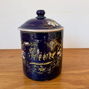 May include: A deep blue ceramic canister with a matching lid, featuring hand-painted gold floral motifs and cursive script text that reads Sucre. The cylindrical container has a glossy finish and a speckled, light-coloured rim on the lid.