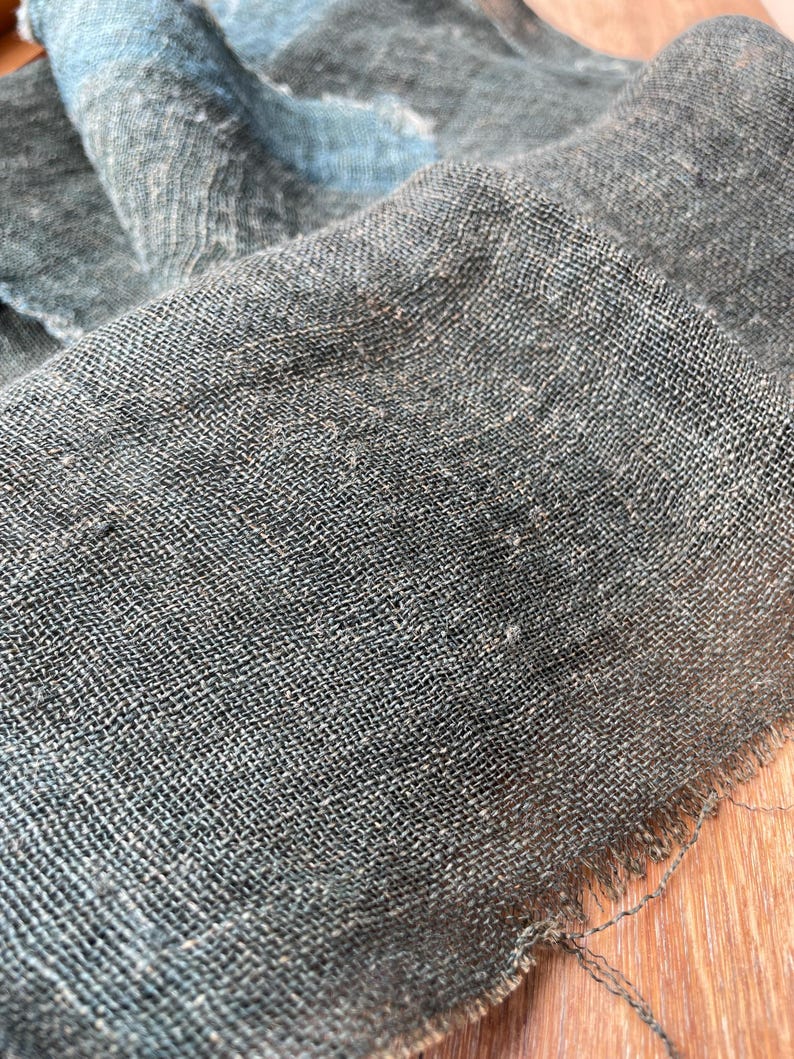 May include: Close-up of a textured, dark gray fabric with a woven pattern. The fabric has a slightly frayed edge and appears to be a natural fiber. Hints of blue are visible in the folds of the material.
