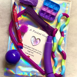May include: A clear plastic bag filled with various sensory toys, including a purple pop-it fidget toy, a purple stretchy tube, a green and blue stretchy snake, a purple mesh ball, a purple stress ball, a purple and green stretchy tube, and a purple candy. A card with the text "A Pocket Hug" and a message of love is included.