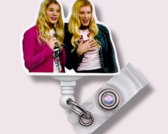 May include: A white badge reel featuring a cartoon image of two women with blonde hair, wearing a pink jacket and a denim jacket. The badge reel has a clear plastic strap and a metal clip.