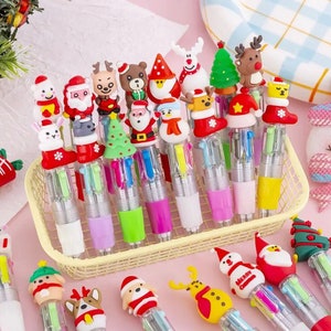 Pu&ograve; includere: Una collezione di penne multicolori colorate con topper a tema natalizio. I topper includono Babbo Natale, renne, pupazzi di neve e alberi di Natale. Le penne sono in un cestino di filo giallo.