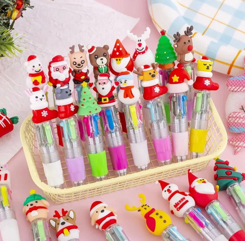 Pu&ograve; includere: Una collezione di penne multicolori colorate con topper a tema natalizio. I topper includono Babbo Natale, renne, pupazzi di neve e alberi di Natale. Le penne sono in un cestino di filo giallo.
