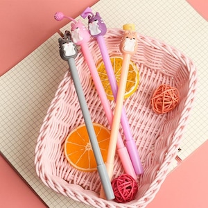 May include: Four colorful pens with cute animal toppers in a pink wicker basket. The pens are gray, pink, purple, and orange. The animal toppers are a cat, a squirrel, a rabbit, and a bear.