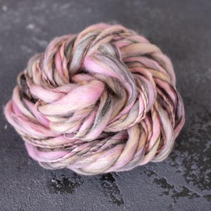 May include: A ball of hand-dyed yarn in shades of pink, gray, and beige. The yarn is tightly wound, showcasing the color variations. Ideal for knitting, crocheting, or other fiber arts projects.