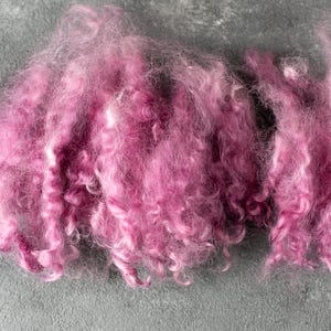May include: A pile of soft, pink fiber, possibly wool or a similar material, rests on a gray surface. The fiber has a fluffy, textured appearance with varying shades of pink, creating a visually appealing display. Ideal for crafting.