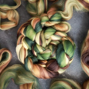 Pode incluir: Um close-up de um novelo de fio colorido em tons de verde, marrom e creme. O fio está disposto em forma circular, com as pontas do fio se estendendo para os lados.