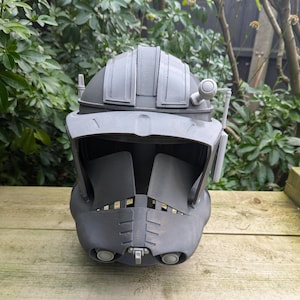 Puede incluir: Un casco de plástico negro y gris con una visera y una pequeña antena en la parte superior. El casco tiene una superficie texturizada y parece ser una réplica de un personaje de una película de ciencia ficción.