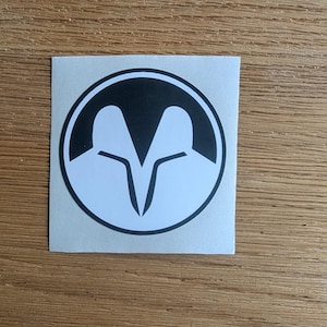 May include: A square sticker featuring a black and white graphic design. The design is a stylized emblem within a black circle, with two white shapes meeting in the center. The sticker is on a light gray backing.