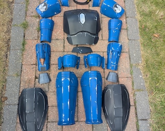 Puede incluir: Un conjunto desmontado de piezas de armadura azules y negras, dispuestas sobre una superficie de ladrillo. La armadura incluye petos, hombreras, protectores de brazos y perneras. Algunas piezas tienen un emblema blanco. La armadura parece estar hecha de plástico o material similar.