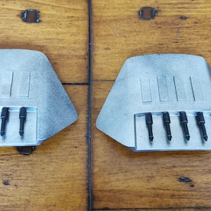 May include: Two silver-colored, angular metal objects with a series of black, cylindrical components. Each piece has a rectangular section with four black cylinders. The objects are on a wooden surface.