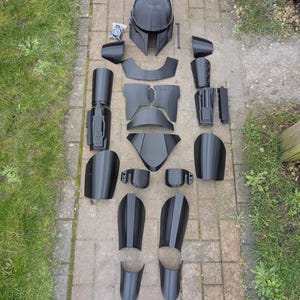 Op de afbeelding: Een gedemonteerde zwarte Mandalorian armor set, uitgestald op een bakstenen oppervlak. De set bevat een helm, borstplaten, schouderstukken, armbeschermers, beenpantser en andere componenten, allemaal in een matte zwarte afwerking. De stukken zijn zo gerangschikt dat ze op een menselijke vorm lijken.