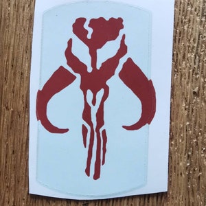 Pode incluir: Um adesivo vermelho e branco com o símbolo mandaloriano. O símbolo é uma caveira estilizada com chifres e uma linha vertical no centro.