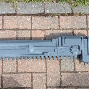 May include: A gray, plastic, saw-like weapon with a pointed tip and a serrated edge. The weapon has a handle with a trigger and a small, round button.