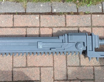 May include: A grey, plastic, saw-like weapon with a pointed tip and a serrated edge. The weapon has a handle with a trigger and a small, round button.