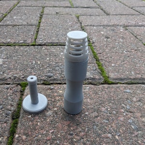 Puede incluir: Dos objetos grises impresos en 3D se muestran sobre una superficie de ladrillo. Uno es de forma cilíndrica con una parte superior ventilada, y el otro es una clavija pequeña de base redonda. Los objetos parecen ser componentes de un artículo más grande.