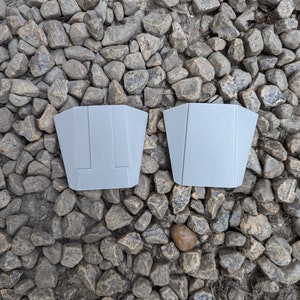 May include: Two gray plastic pieces with a geometric design. The pieces are on a background of small gray rocks.
