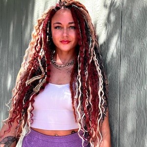 May include: A white ribbed crop top and purple athletic shorts paired with long, multi-textured hair extensions in shades of deep red, black, and cream. The hair features various twists, braids, and decorative wraps throughout the long, wavy strands.