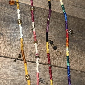 May include: Four hair wraps made of multicolored thread wound around strands. The wraps feature various metallic beads, small charms including keys and a heart, and circular pendants. The threads transition through shades of yellow, white, red, purple, black, and rainbow colors, creating a vibrant and textured appearance for hair styling.