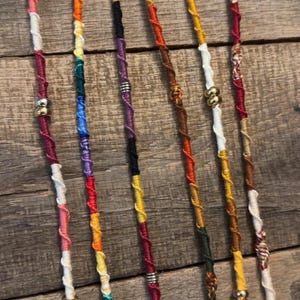 Puede incluir: Seis extensiones de cabello largas hechas de hilo multicolor enrollado alrededor de un núcleo. Cada una presenta una combinación única de hilos rojos, amarillos, azules, verdes, blancos y marrones, con pequeñas cuentas metálicas plateadas y doradas.