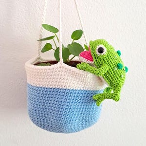 May include: A blue and white crocheted hanging planter with a green crocheted frog hanging on the side. The planter is filled with a small green plant.