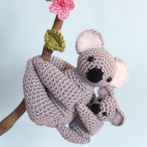May include: A gray crocheted koala holding a smaller gray crocheted koala. The koalas are hanging from a branch with a pink crocheted flower and green leaves.