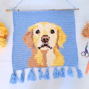 May include: Crocheted wall hanging featuring a golden retriever dog with blue tassels. The hanging is made with light blue, yellow, and brown yarn.