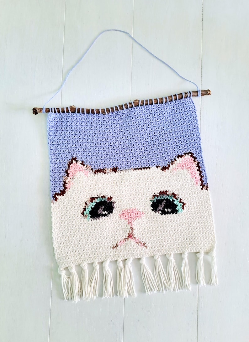 May include: Crocheted wall hanging featuring a white cat face with pink ears, nose, and mouth, and blue eyes. The cat face is on a purple background with white fringe at the bottom.