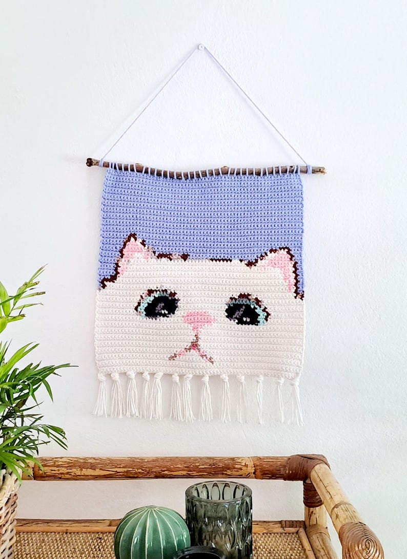 May include: A crocheted wall hanging featuring a white cat face with blue eyes and a pink nose. The cat face is on a white background with a purple background above it. The hanging has white tassels at the bottom and is hung on a wooden stick.