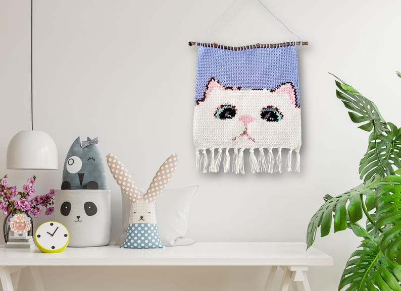 May include: A white crocheted wall hanging with a cat face design. The cat has pink cheeks and blue eyes. The hanging has a light blue background and white fringe.