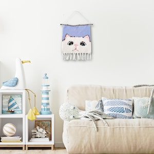 May include: A white cat face woven wall hanging with a light blue background. The cat has pink ears and a pink nose. The hanging has a fringe border.