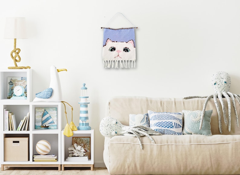 May include: A white cat face woven wall hanging with a light blue background. The cat has pink ears and a pink nose. The hanging has a fringe border.