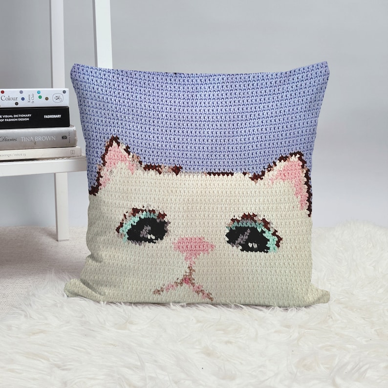 May include: A crocheted pillow with a white cat face design. The cat has blue eyes and a pink nose. The background of the pillow is a light purple.