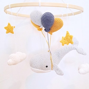 Peut inclure: Un mobile en crochet en forme de baleine avec des ballons, des nuages et des étoiles gris, blancs et jaunes suspendus à un anneau en bois.