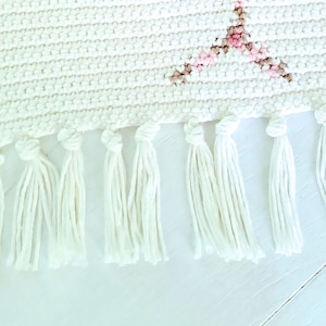 May include: White woven wall hanging with a pink and brown cat face design. The hanging has white fringe along the bottom.