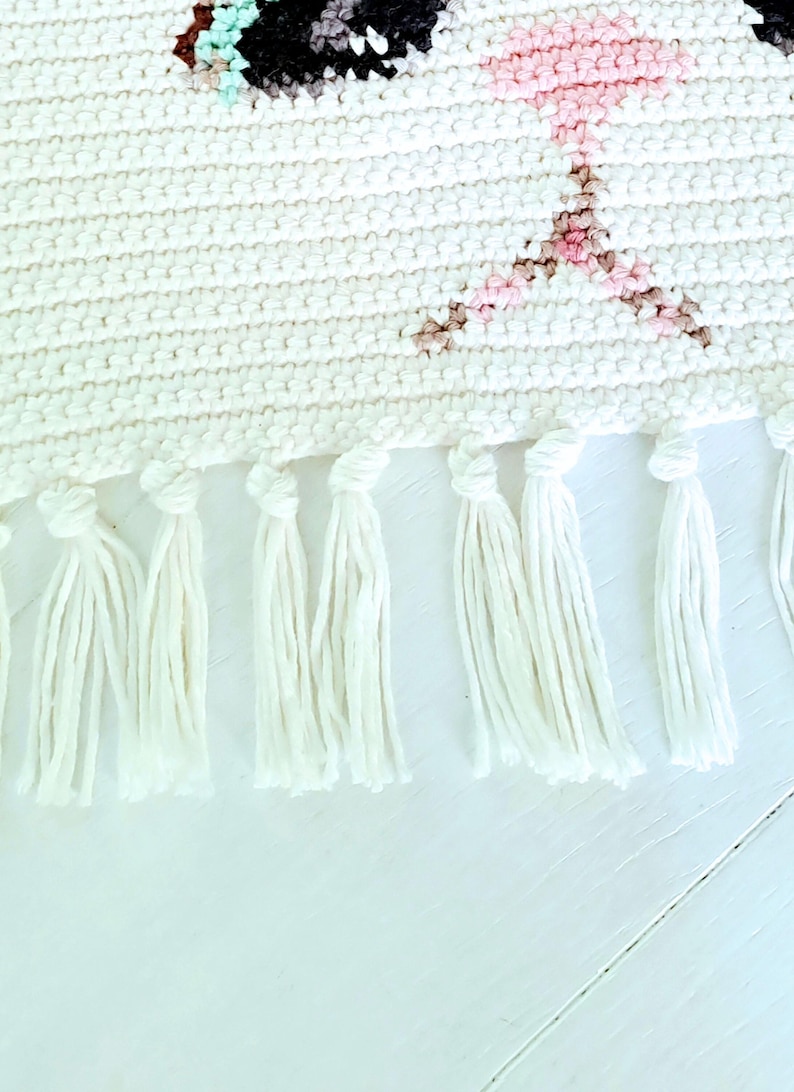 May include: White woven wall hanging with a pink and brown cat face design. The hanging has white fringe along the bottom.