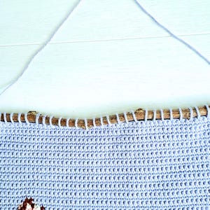 May include: A light blue crocheted wall hanging with a brown branch at the top. The hanging features a white and pink design of a cat's face.
