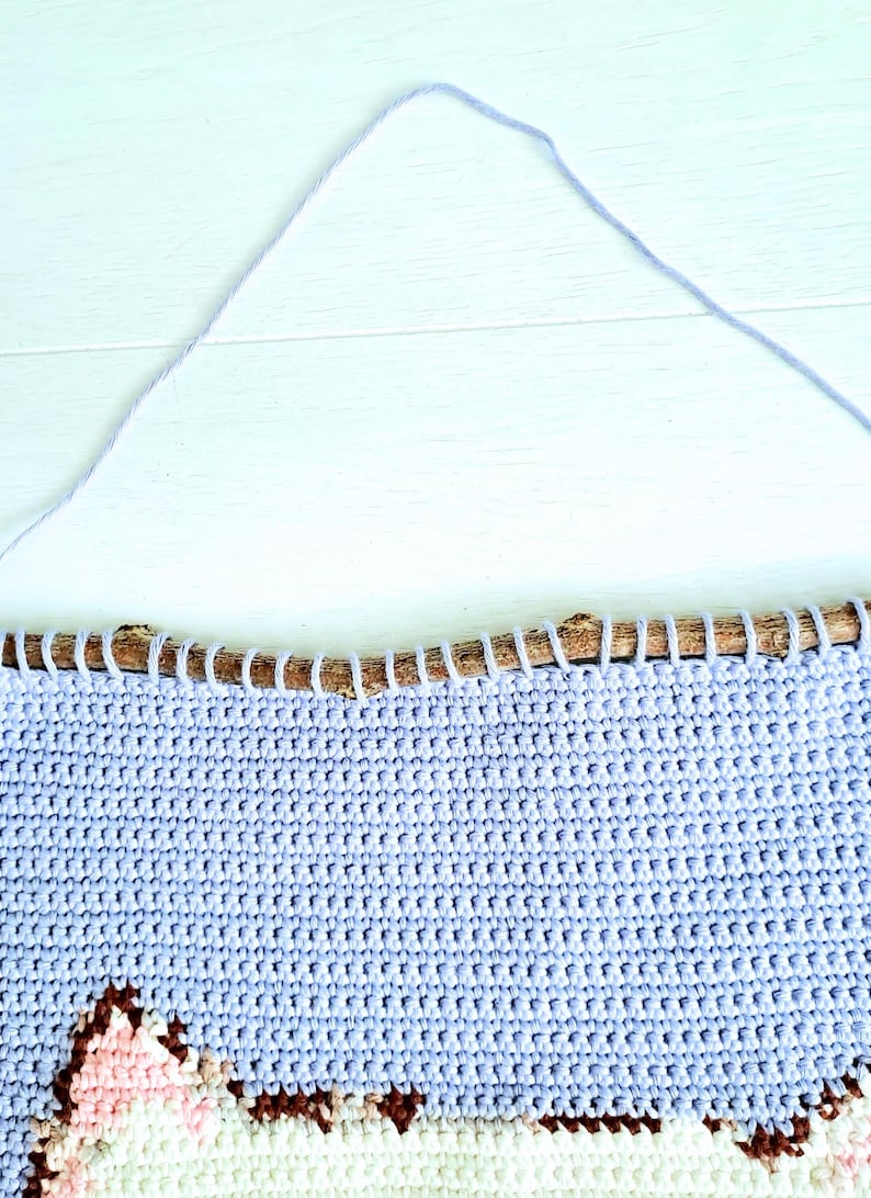 May include: A light blue crocheted wall hanging with a brown branch at the top. The hanging features a white and pink design of a cat's face.
