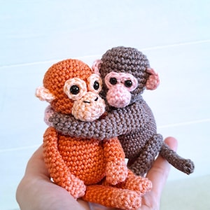 Puede incluir: Dos monos de ganchillo hechos a mano en colores cálidos. Uno es naranja, el otro marrón, y se abrazan. Los monos tienen acentos rosas en las orejas y la cara. Los juguetes son pequeños y parecen suaves.