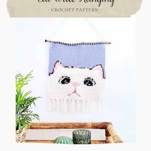 May include: Crochet cat wall hanging with a light blue and white color scheme. The cat has black eyes and a pink nose. The hanging is attached to a wooden stick with a white string. The text "Cat Wall Hanging - CROCHET PATTERN -" is at the top of the image. The text "BIRDSANDCRICKETS" is at the bottom of the image.