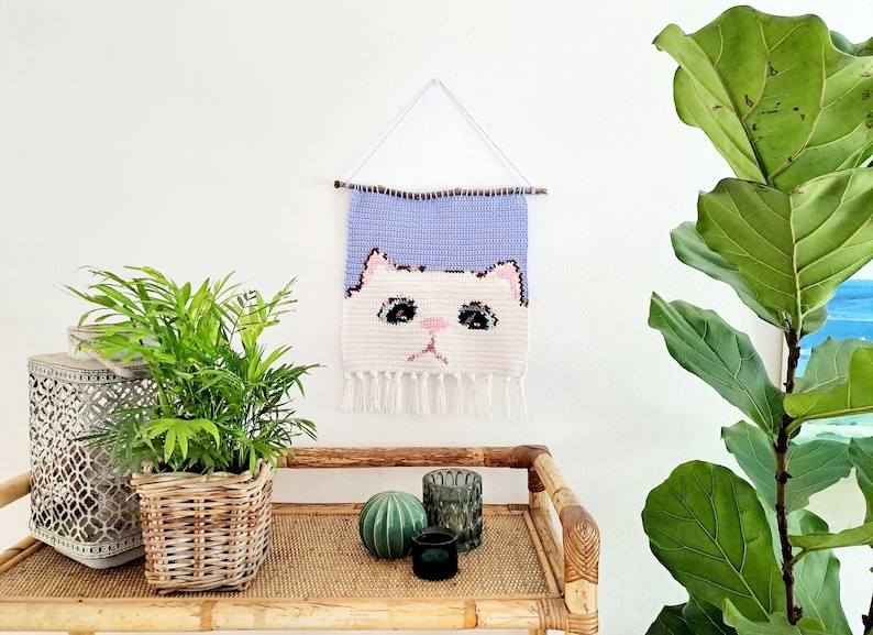 May include: A crocheted wall hanging featuring a white cat face with blue eyes and a pink nose. The cat face is on a light purple background with white fringe at the bottom. The wall hanging is displayed on a white wall with a wooden stick at the top.